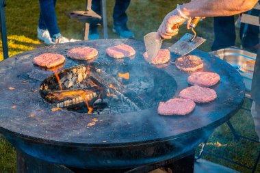 Çimenlikte, büyük yuvarlak ahşap ızgara hamburger için köfte kızartıyorlar. Aşçı kızarmış pirzolaları çevirir. Şehir parkında barbekü festivali. Sokak Fast food.