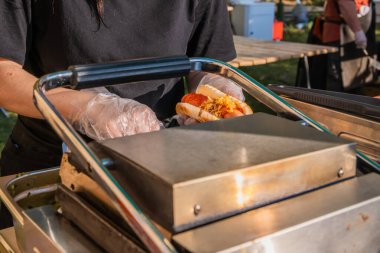 Bir şefin eldivenli elleri yeni kızartılmış bir sosisliyi dolduruyor. Şehir parkında barbekü festivali. Sokak Fast food.