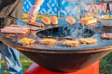 Çimenlikte, büyük yuvarlak ahşap ızgara hamburger için köfte kızartıyorlar. Aşçı kızarmış pirzolaları çevirir. Şehir parkında barbekü festivali. Sokak Fast food.