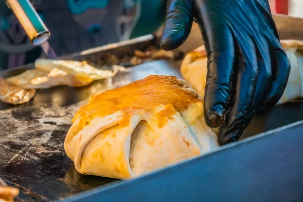 Mexican food, a steak burger wrapped in a tortilla under melted cheese on the grill of an outdoor restaurant. Food festival in the city park. Street fast food.