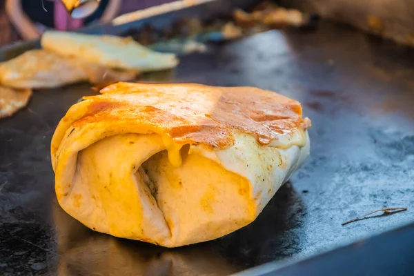 Mexican food, a steak burger wrapped in a tortilla under melted cheese on the grill of an outdoor restaurant. Food festival in the city park. Street fast food.