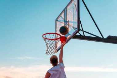 Genç bir basketbolcu topu mavi gökyüzüne doğru basket sepetine atıyor. Açık alanda şeffaf plastik basketbol kalkanı..