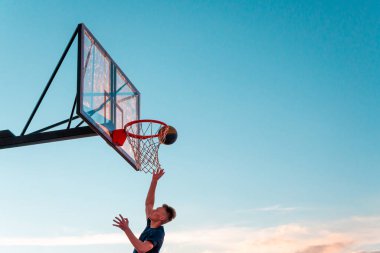 Genç bir basketbolcu topu mavi gökyüzüne doğru basket sepetine atıyor. Açık alanda şeffaf plastik basketbol kalkanı..