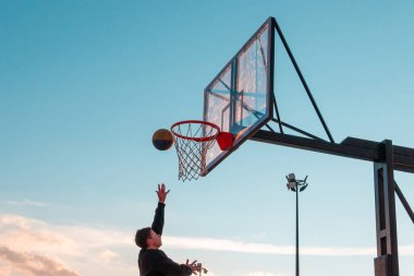 Genç bir basketbolcu topu mavi gökyüzüne doğru basket sepetine atıyor. Açık alanda şeffaf plastik basketbol kalkanı..