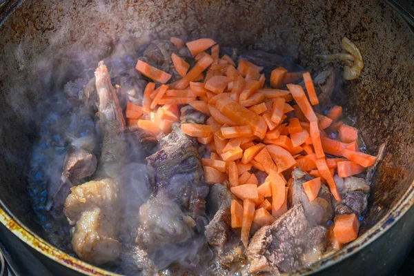 Sliced carrots and pieces of meat are fried in the process of cooking pilaf in boiling oil in a cauldron on a traditional Asian oven. Step-by-step recipe for real pilaf.