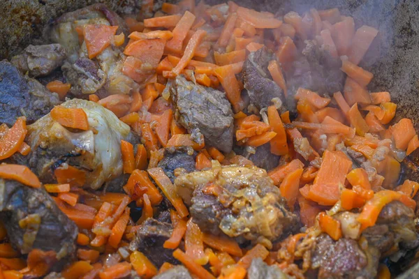 Sliced carrots and pieces of meat in a cauldron in the process of cooking pilaf on a traditional Asian oven. Step-by-step recipe for real pilaf.