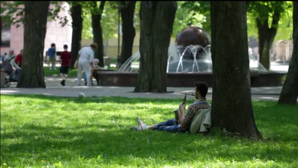 Молодий чоловік грає на саксофоні, сидячи під деревом у парку — стокове відео