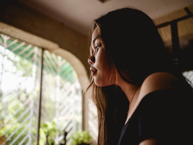 Depresyondaki genç Asyalı bir kadın, bir odada yalnız ve umutsuz bir bakışla somurtuyor. Pencereden gelen hafif doğal ışık. Depresyon kavramı, kalp kırıklığı.