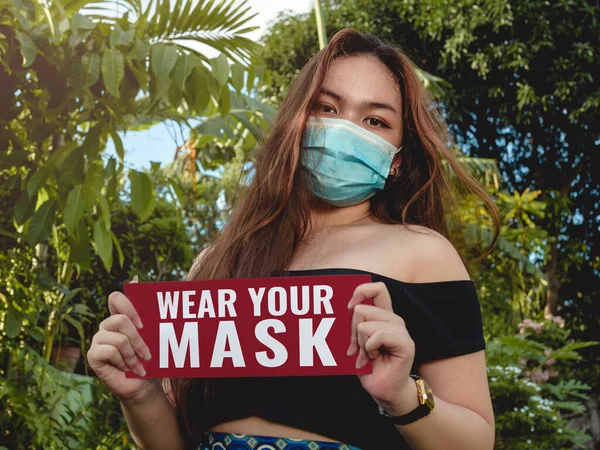 若いアジアの女の子は屋外であなたのマスクを着用するためのフレンドリーなリマインダとサインを保持しています — ストック写真