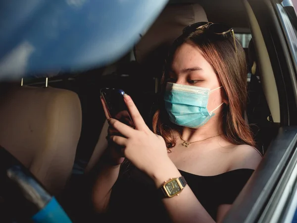 ガレージや駐車場や道路脇に駐車している車の中で 外科用マスクをした若いアジアの女の子がソーシャルメディアでチャット中です — ストック写真