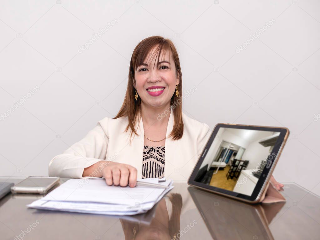 A successful asian realtor with document papers and a tablet showing a portfolio of her properties for sale.