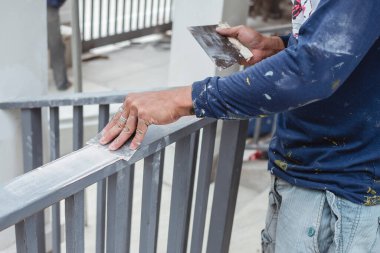 A worker applies epoxy putty to smooth a steel railing by the porch of a home under construction. Closeup of hand holding putty knife. clipart