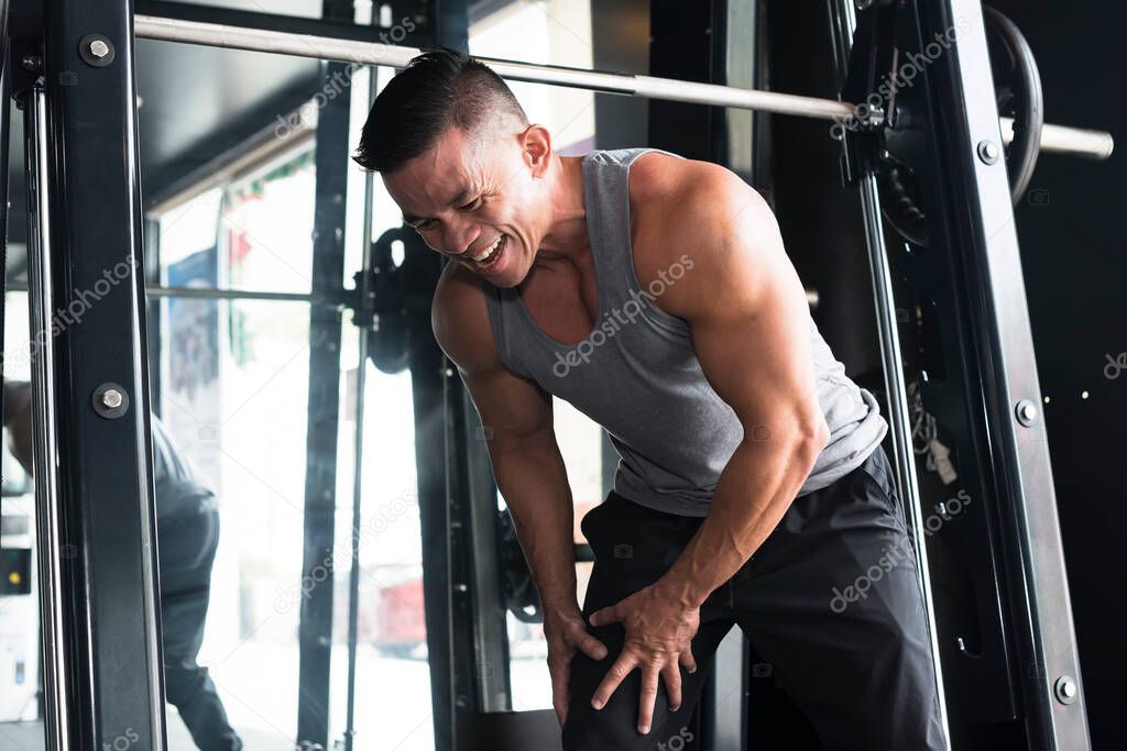 A asian man experiences leg cramping and grimaces in pain. He tightly clutches his thighs. By the Smith machine at a gym.