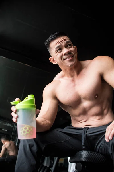 A fit and handsome middle aged male sitting on a bench and holding a sports drink after a vigorous workout at the gym. In his early 40s but very youthful looking.
