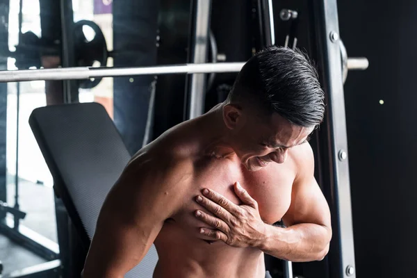 An asian male suffers a torn pectoral muscle while doing heavy incline bench presses on a Smith Machine at a gym or fitness center. A common workout injury