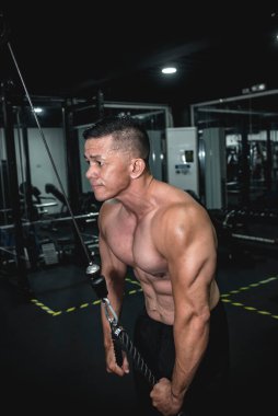 Odaklanmış ve kaslı bir adam trisepslerini salonda halatla iterek eğitiyor. Kol çalışması.