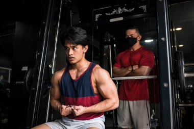 Genç ve yakışıklı Asyalı bir adam kaslarını esnetirken maskeli bir eğitmen şu ana kadar ilerlemeyi ve kas gelişimini inceliyor..