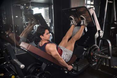 Yakışıklı genç bir sporcu bacak presi makinesinde bacak kaslarını çalıştırırken diğer aynada vücudunu ve görünüşünü kontrol eder..