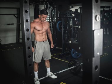 Tişörtsüz ve ciddi görünümlü Asyalı bir adam yataklı zemindeki halterlere bakıyor. Spor salonunda..