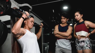 Bir fitness eğitmeni, spor salonundaki iki müşteriye omuz presi makinesinin nasıl kullanılacağını düzgün bir şekilde gösterir..