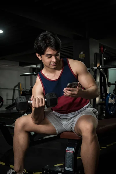A handsome young asian man watches a video on his phone while working out, training biceps. Distracted or multitasking.