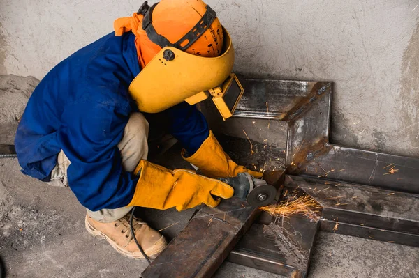 A man in full protective gear cuts diagonally into a steel rectangular tube with an angle grinder. At a construction