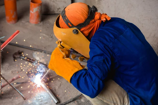 A man crouched and in full welding gear welds together a makeshift trowel, putty knife or scraper from two pieces of metal. Ingenuity and resourcefulness at the construction site.