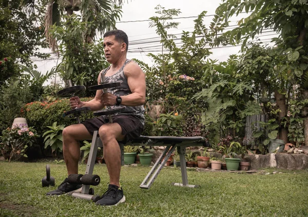 A fit middle aged asian guy prepares to lift seated dumbbell shoulder presses on a bench at his front yard. Fitness and working out at home.