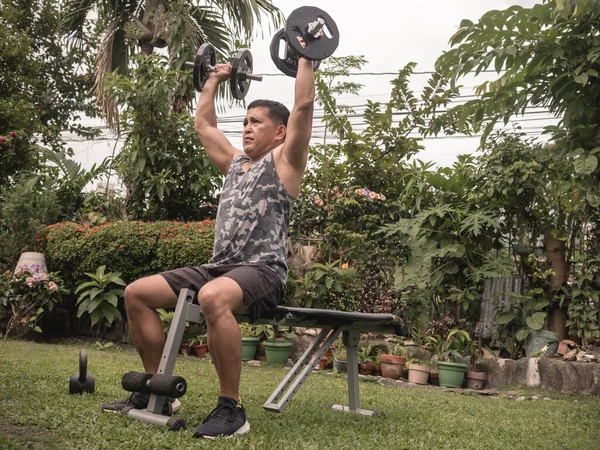 A fit and athletic middle aged asian guy does seated dumbbell shoulder presses on a bench at his front yard. Fitness and working out at home.