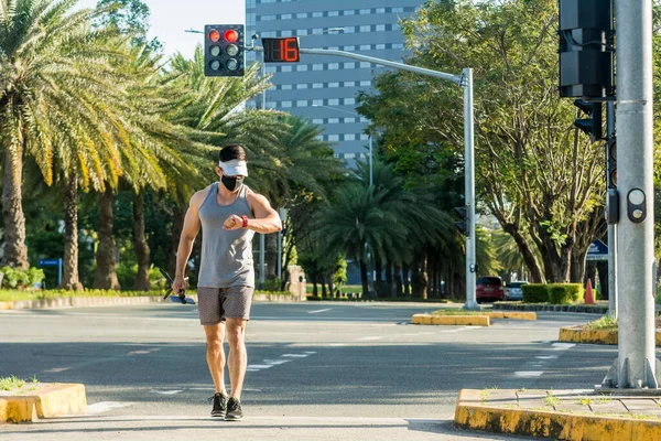 A fit asian guy in sportswear checks the time while walking around the city for fitness and health purposes. Wearing a face mask, new normal scene outdoors.