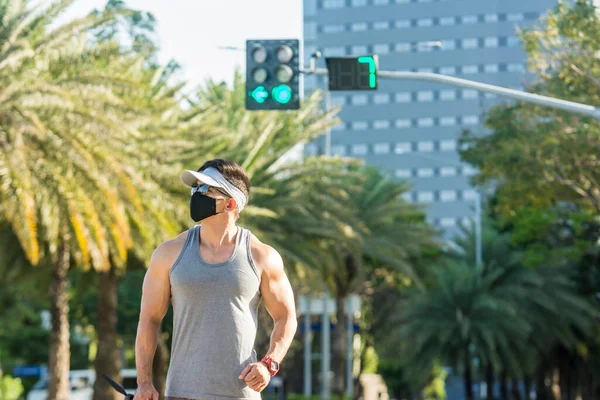 A buff asian guy walks at a beautiful city street lined with date palms. Wearing a face mask, new normal scene outdoors.