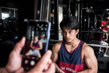 Genç bir fitness modeli kollarını esnetiyor ve spor salonundaki ilerlemesini izlemek için fotoğrafını çekiyor. Fiziksel gelişim ve kas kazanımları belgeleniyor.