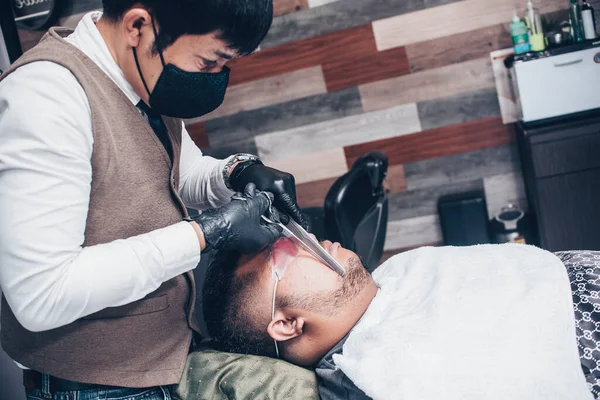 Азиатский Парикмахер Маске Лица Обрезает Бакенбарды Своего Клиента Ножницами Расчёской — стоковое фото