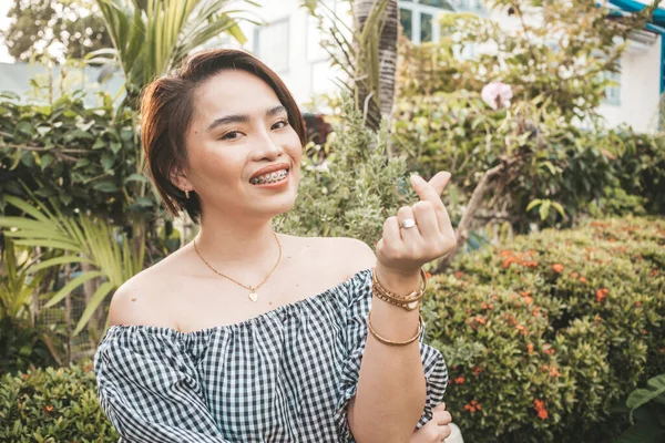 A beautiful short haired asian woman wearing an off shoulder top gives a finger heart gesture. Summer outfit.