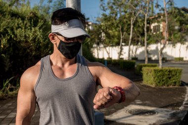 Yüz maskesi takmış fit bir Asyalı saati kontrol eder, ya da nabız, kalp atışı, mesafe ya da kalori gibi verileri. Açık hava sahnesi.