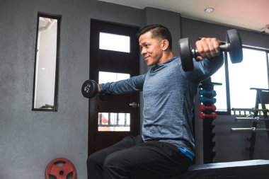 Tişörtlü, fit bir Asyalı yan yana oturmuş zam yapıyor. Omuz egzersizi ve antrenmanı. Spor salonu ayarlarını aç.