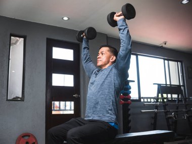 Sweatshirt giyen atletik bir adam dambıl ütüler. Omuz egzersizi ve antrenmanı. Spor salonu ayarlarını aç.