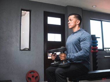 Tişörtlü formalı bir adam omuzlarında iki dambıl, dizlerinde dinlenerek egzersiz yapmaya hazırlanıyor. Spor salonu ayarlarını aç.