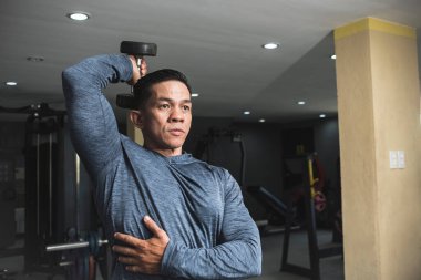 Tişörtlü, formda, atletik Asyalı bir adam, bir kol uzatması için halterde oturuyor. Kol kaslarını çalıştırmak ve çalıştırmak. Spor salonu ayarlarını aç.