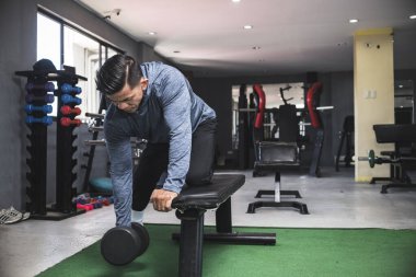 Tişörtlü kaslı ve kaslı bir adam tek koluyla dambıl gibi kürek çekiyor. Sırt egzersizi. Spor salonunda..