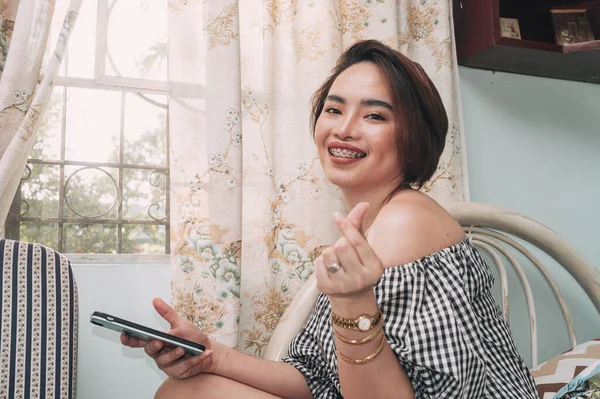 A short haired woman wearing a off shoulder top gives a finger heart sign while checking her social media inside the bedroom.