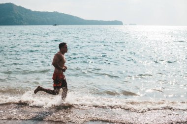 Formda ve atletik bir erkek kıyı şeridi boyunca yalın ayak koşuyor. Manzaralı bir plajda eğlence ve düşük yoğunluklu kardiyo..