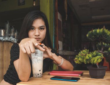 Üzgün genç Asyalı bir kadın, bir kafede sütlü çay yudumlarken küçümseyici bir bakışla birini işaret eder..