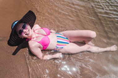 Deniz kıyısında kumların üzerinde uzanmış, koyu güneş şapkası takmış ve ışıl ışıl gülümseyen bir kadın. Seyahat ve tatil plaj konseptinde..