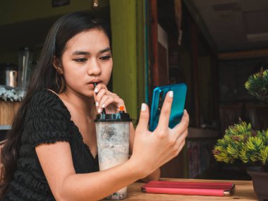 Genç bir Filipinli kadın küçük bir kafede sütlü çay yudumlarken sosyal medyasını kontrol etmekle meşgul. Olası bağımlılık, aşırı kullanım ya da saplantı.