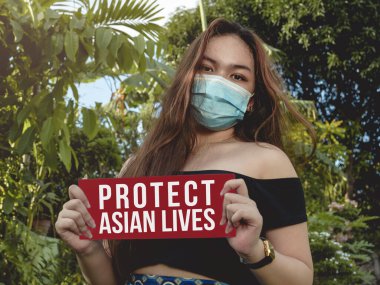 Yüz maskesi takan Filipinli Amerikalı genç bir kadın dışarıda Asyalıları koruyan bir tabela tutuyor.