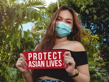 A young Filipino-American woman wearing a face mask holds a protect asian lives sign outdoors clipart