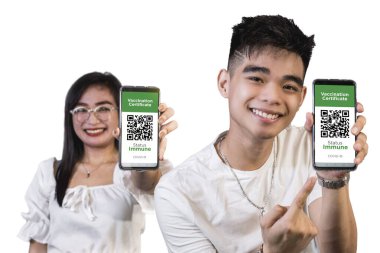 İki genç Asyalı dijital dokunulmazlık pasaportlarını veya aşı kanıtlarını telefonlarında uygulama olarak sunuyorlar. Düz beyaz arka plan. Telefona odaklanmış.