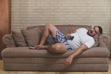 An overweight, unshaven and lazy man of mixed race sleeps on the couch, sprawled out and still holding his cellphone. clipart