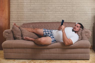 Tipik bir kanepe patatesi - iç çamaşırlı bir adam kanepede uzanıyor ve video izliyor, mobil oyunlar oynuyor veya sosyal medyada.